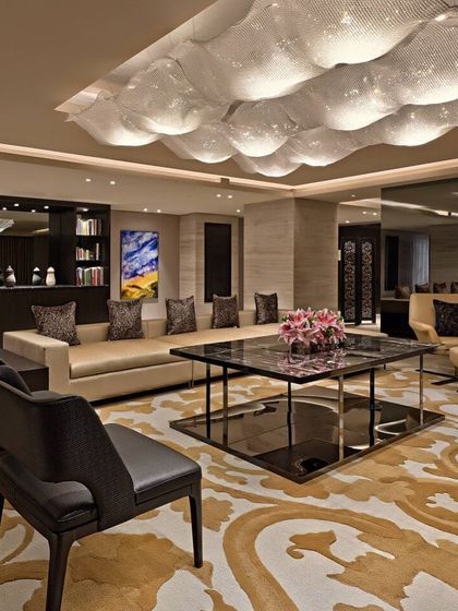 A luxurious hotel suite living area. The design features a unique, cloud-like ceiling light installation, a custom patterned rug, and a curated selection of contemporary furniture and art.