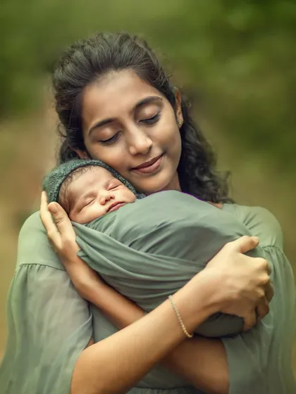 A new mother, with her eyes gently closed, cradles her sleeping, swaddled newborn against a soft, green forest backdrop. This outdoor newborn portrait captures the pure serenity and overwhelming love of early motherhood.
