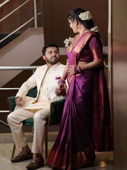 A royal and romantic pose. The groom is seated while his bride stands, showcasing her elegant purple saree and coordinated makeup.