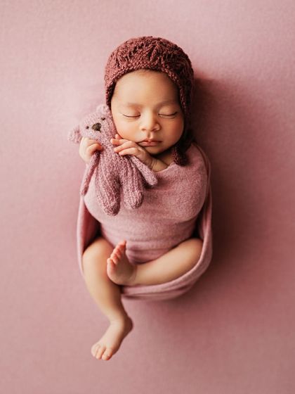 A full-body shot of a baby wrapped in pink, holding a tiny knitted teddy bear. This pose is called the "potato sack" and is perfectly safe and comfortable for newborns.