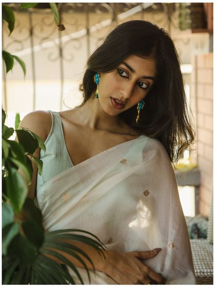 A soft and elegant portrait using natural light indoors. The plants in the foreground create a beautiful, natural frame, adding depth to the image. The model's off-camera gaze gives the shot a candid, story-driven feel.