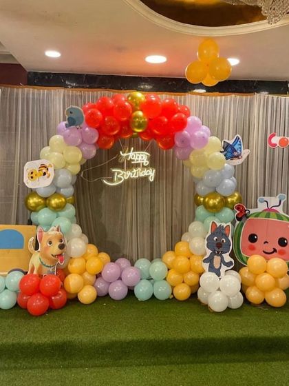 A Cocomelon themed circular balloon arch on a green mat, perfect for a cake-cutting ceremony. The setup includes character cutouts and a 'Happy Birthday' neon sign.