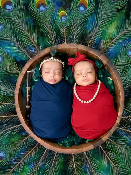 Double the love, double the charm. These precious twins are styled as little Radha and Krishna, nestled in a bowl against a backdrop of peacock feathers. Capturing twins is a special art, and this image shows the divine bond they share from the very beginning.