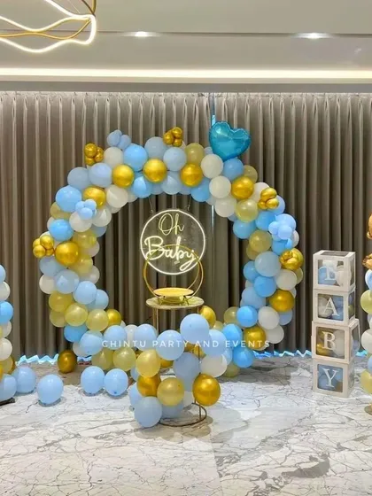 An elegant baby shower decoration for a home setting. This setup includes a full balloon ring in blue, white, and gold, with an 'Oh Baby' neon sign, 'BABY' blocks, and balloon columns topped with themed foil balloons.