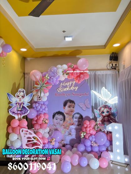 A magical fairy-themed first birthday for Radhya. This setup includes a custom photo backdrop, fairy character cutouts, and a pink and purple balloon arrangement with an illuminated number one.
