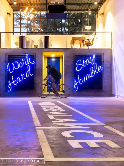 We transformed an old warehouse into an energetic office space, using neon signs like 'Work Hard Stay Humble' to motivate and inspire. The open-plan design includes unique features like an indoor cycling track, blending industrial grit with a playful, modern spirit to create a workspace that's anything but ordinary.