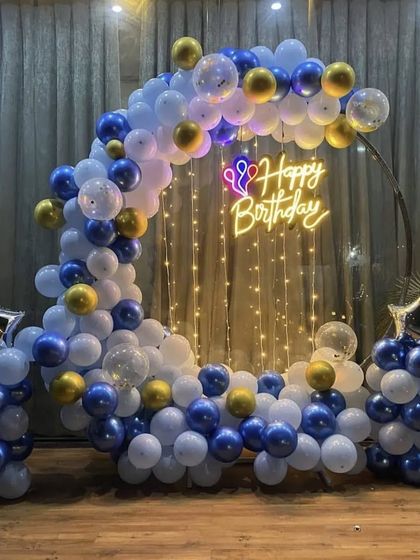 A classic blue, white, and gold balloon ring backdrop for a birthday, illuminated by fairy lights and a central neon sign.