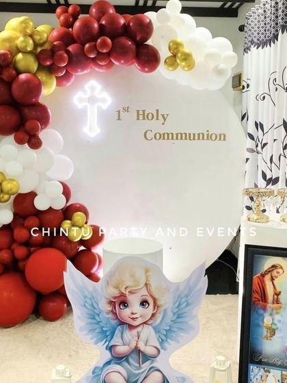 A First Holy Communion decoration for a girl with an angel theme. The setup uses a red, white, and gold balloon arch and a cute angel cutout.