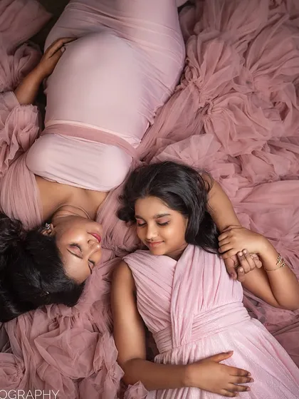 In a beautifully styled studio setting, a mother-to-be and her daughter lie together on a bed of soft pink tulle, sharing a quiet, loving moment. This image highlights the tender bond between a mother and her child as they await a new arrival.