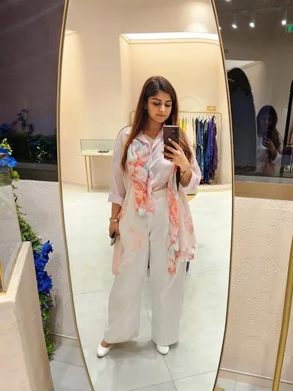 A mirror selfie capturing a soft, professional look. The combination of a pale pink shirt with a floral scarf and white trousers creates an elegant and approachable vibe. This is how you wear pink to stand out gracefully.