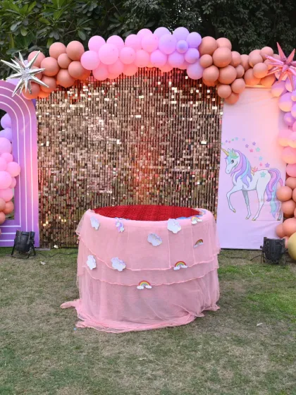 A magical unicorn-themed birthday setup in an outdoor garden. The decor features a gold sequin backdrop, a rainbow arch, and a special tulle-draped table for the cake.