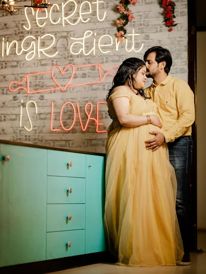 An intimate couple's moment captured in a modern indoor setting. The warm glow of the neon sign, "secret ingredient is LOVE," adds a fun, contemporary vibe to this maternity portrait.
