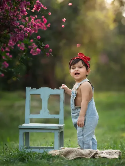 A toddler in denim overalls stands by a small chair under a tree with pink blossoms. A vibrant and happy outdoor portrait.