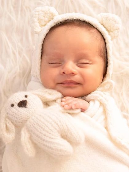 An overhead shot of a smiling baby in a bear bonnet, cuddled up with a toy on a fluffy white blanket. This simple setup is all about warmth and comfort.