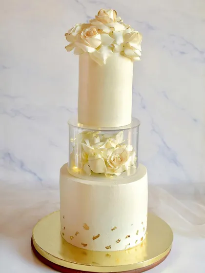 A classic and elegant three-tier wedding cake with a transparent spacer. The design is kept clean and timeless in all-white, decorated with beautiful fresh white roses and touches of gold leaf.