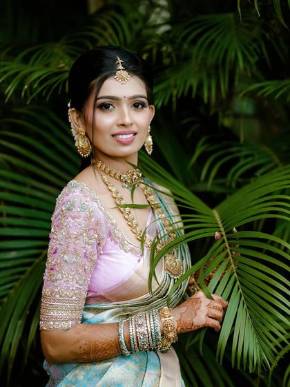 A smiling bride holds a palm leaf, her pastel saree and embroidered blouse looking fresh and vibrant against the green background.