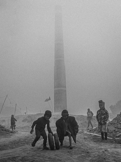 Children play with a tire in the foggy, industrial landscape of a brick kiln, a poignant black and white image of childhood resilience.