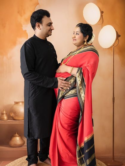 An intimate moment between a couple in a warmly lit studio setting with traditional Indian decor. Their hands gently cradle the baby bump, showing their shared love.