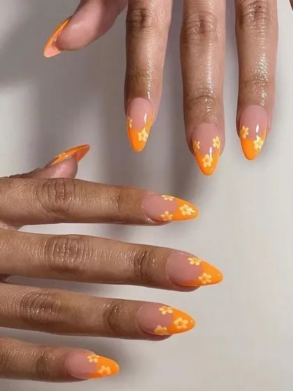 Pointed almond nails with a negative space design, featuring bright orange tips decorated with small, simple white flowers. A fresh and fun take on a floral manicure.