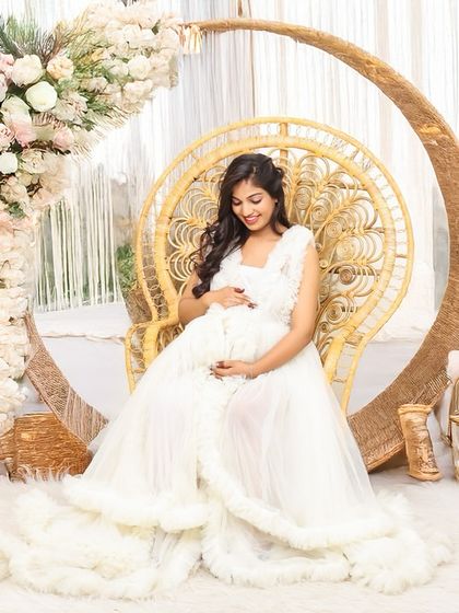 A serene mother-to-be in a white ruffled gown, seated on a peacock chair within a floral and wicker arch. This studio setup creates a dreamy, light, and airy feel.