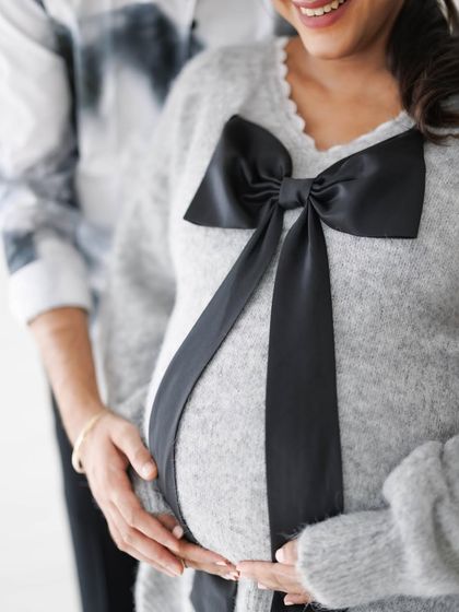 A close-up on the baby bump, adorned with a large bow, with the partner's hands gently holding her. A symbol of the precious gift they share.