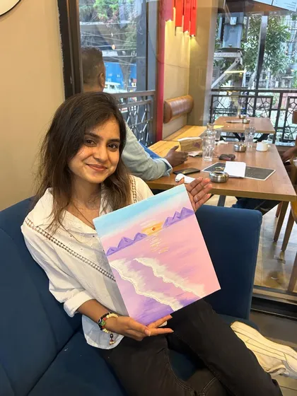 A participant sitting on a blue couch, proudly holding her finished pink beach painting.