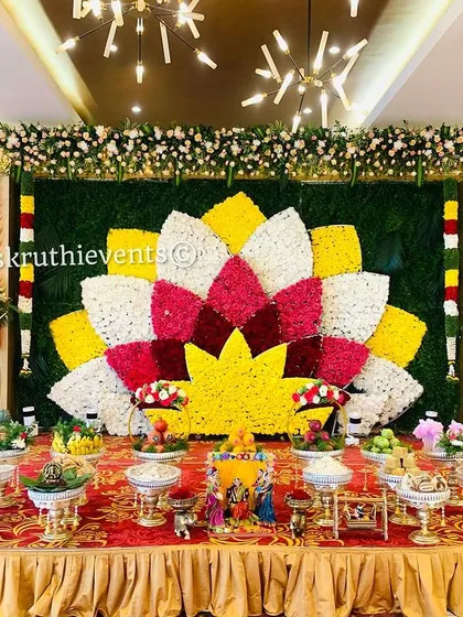 A magnificent lotus-shaped floral backdrop as the centerpiece for a baby shower. The stage is further enhanced by a beautiful arrangement of offerings and traditional dolls, making it a visual delight.