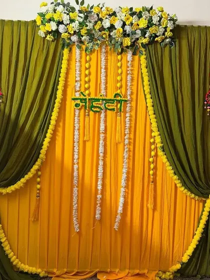 A simple yet elegant Haldi backdrop using green and yellow drapes. The hanging marigold and jasmine flower strings add a traditional touch to this indoor home setup.