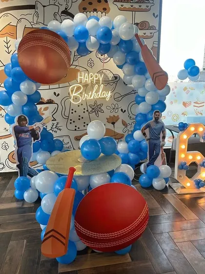 A cricket-themed 6th birthday party for a young sports fan. The setup includes giant cricket bat and ball props, player cutouts, and a blue and white balloon arch.