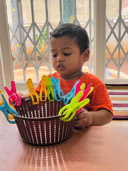 This toddler is developing his pincer grasp and sense of order by clipping clothespins onto a basket. Even simple activities are designed to be purposeful and build foundational skills.