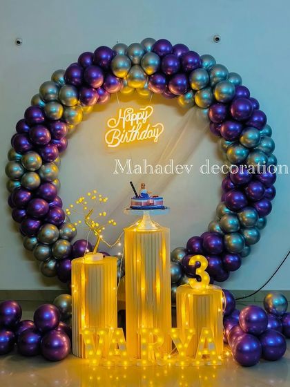 A sophisticated birthday setup with a full circular arch of metallic purple and silver balloons. The decor is enhanced by pedestals wrapped in lights and a 'Happy Birthday' neon sign.
