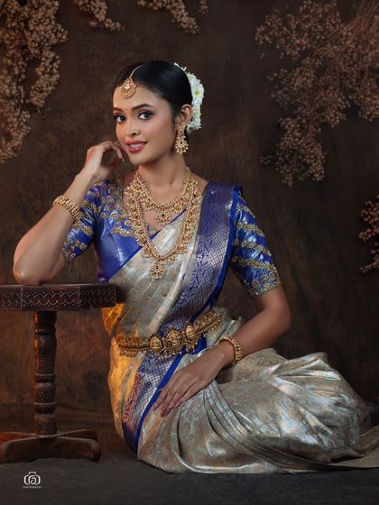 Another elegant seated portrait in a traditional South Indian saree. The pose is relaxed and graceful, with the lighting designed to highlight the rich texture of the silk and gold jewelry.