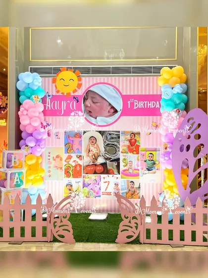 A charming photo milestone board for Aayra's first birthday. The display is set against a pink picket fence with butterfly accents and a cheerful sun cutout, showcasing photos from her first year.