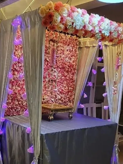 An elegant and sophisticated mandap featuring a dense pink floral wall as the backdrop. The setup is framed with shimmering silver drapes and accented with soft purple lighting, creating a truly special and premium look for your home.