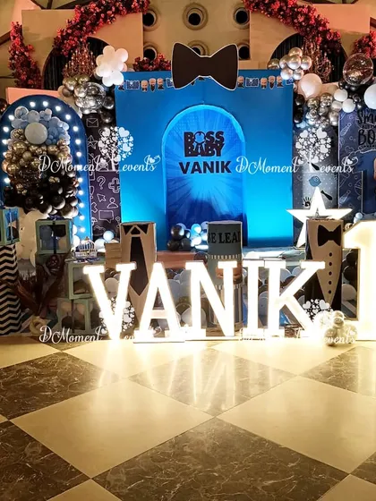 A stylish Boss Baby stage for Vanik's first birthday. The decor features a bold blue backdrop, a large bowtie prop, and marquee letters against a checkered floor.