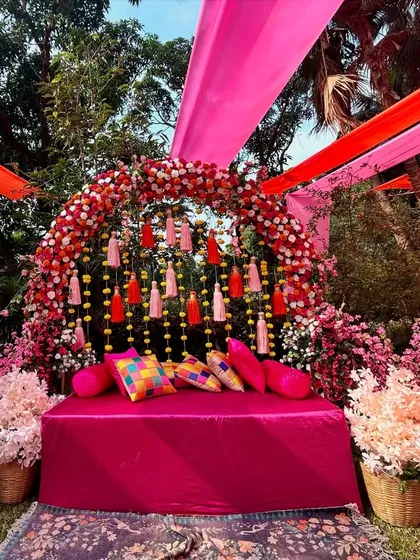 A lush and vibrant Mehendi seating area, featuring a floral arch of pink and red blooms, colorful cushions, and traditional tassels. This setup is perfect for a romantic and festive feel.