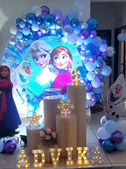 A beautiful Frozen themed birthday setup with a circular backdrop featuring Elsa and Anna, surrounded by a purple and blue balloon garland.
