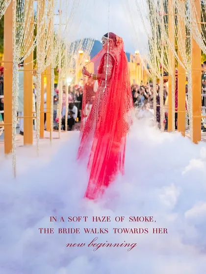 In a soft haze of smoke, the bride walks towards her new beginning. This dramatic entrance shot highlights the beauty of her red lehenga and long veil.