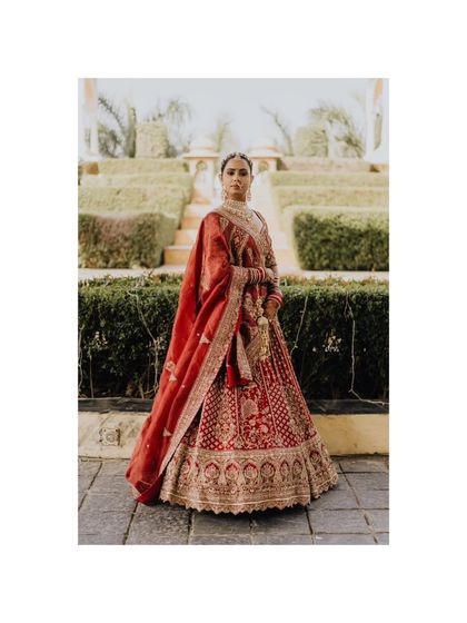 A full-length portrait of the bride, Dhwani, showcasing her exquisite red lehenga against the backdrop of a beautiful garden.