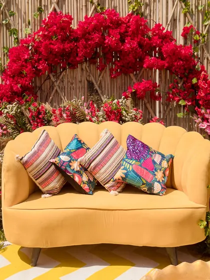 A close-up of the sunshine-yellow sofa, adorned with colorful and patterned cushions, set against the bougainvillea arch.