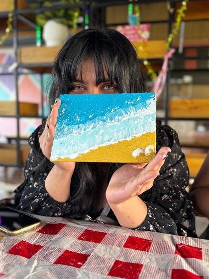 A participant playfully peeks over her finished beach scene texture painting. It's a fun way to escape and create your own little slice of paradise.