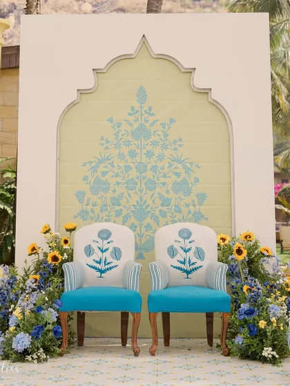 The main seating for the Haldi ceremony, featuring custom-printed chairs set against a backdrop with traditional Indian floral motifs. The blue and yellow color scheme is a modern twist on Haldi decor.