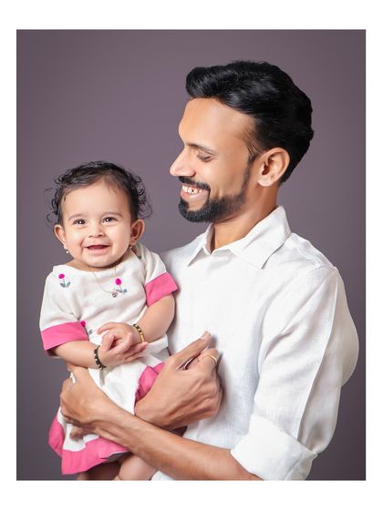 A father holds his baby, both sharing a happy smile. The simple grey background keeps the focus entirely on their joyful connection.