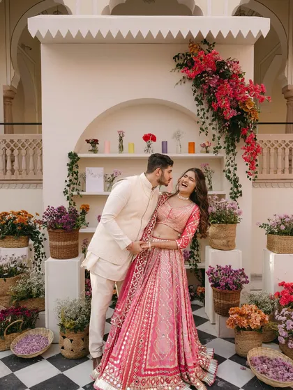 A candid moment of laughter at a colorful Haldi in Jaipur. The couple is framed by a backdrop of shelved niches filled with baskets of fresh flowers, adding texture and life to the setting.