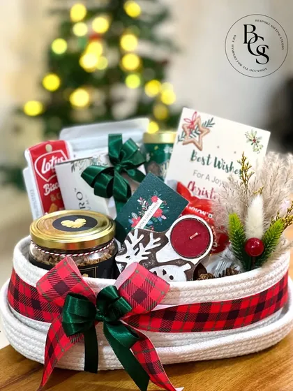 A cozy and festive Christmas hamper arranged in a rope basket with plaid ribbon. Filled with holiday treats like Biscoff cookies, a scented candle, and gingerbread, it’s the perfect gift to place under the tree.