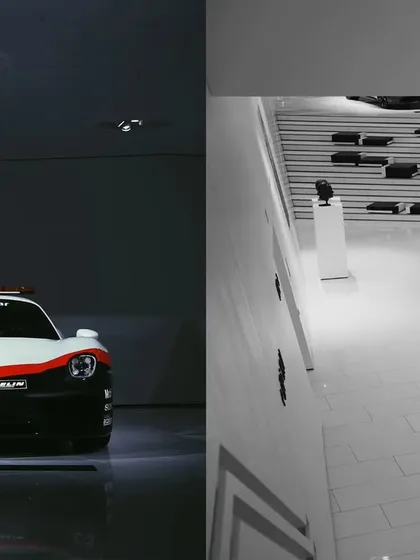 A dramatic, high-contrast shot of a Porsche 911 safety car, using shadows to create a powerful mood. The other half of the image shows the minimalist architecture of the museum, adding context and scale.