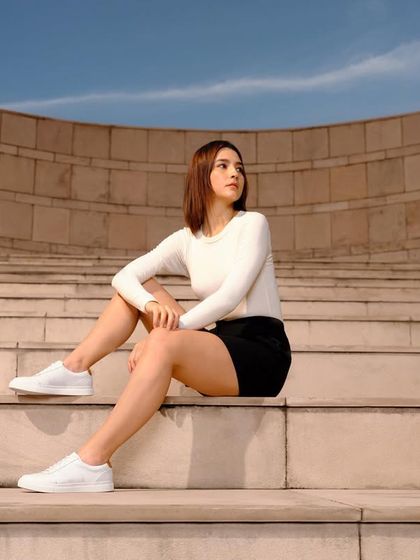 A clean, commercial look for a shoe campaign. The styling is minimalist and classic with a white top, black skirt, and white sneakers, set against a modern architectural background.