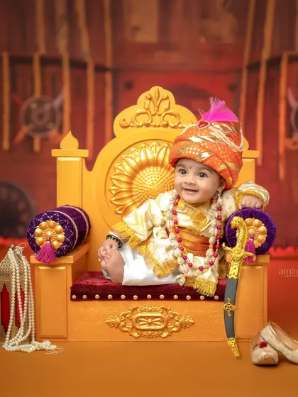 Capturing the spirit of a young king, this Bal Shivba themed portrait features a six-month-old baby in royal attire on a miniature throne. The setup includes a toy sword and traditional shoes, set against a backdrop of a Maratha fort.