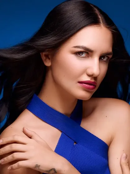 A beauty shot with a focus on healthy, flowing hair against a rich blue background. The model's pose highlights the neckline of her top while keeping the attention on her flawless features.