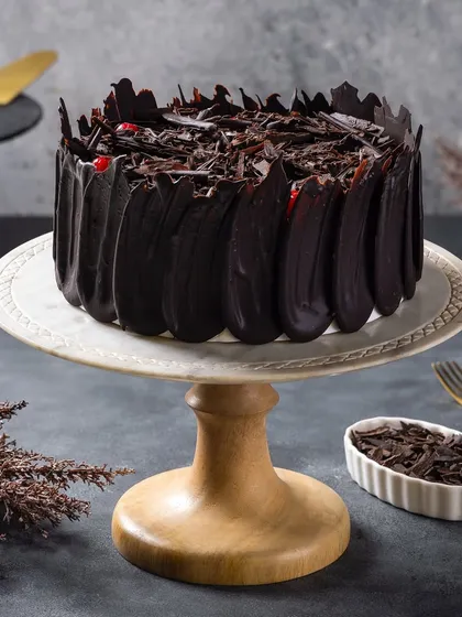 A black forest cake with intricate chocolate shavings. The lighting is designed to catch the glossy finish and sharp edges of the chocolate.
